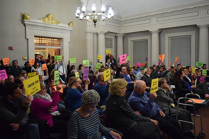 Audience members hold neon orange, yellow, pink and green signs while Mayor Earl Leiken speaks. 