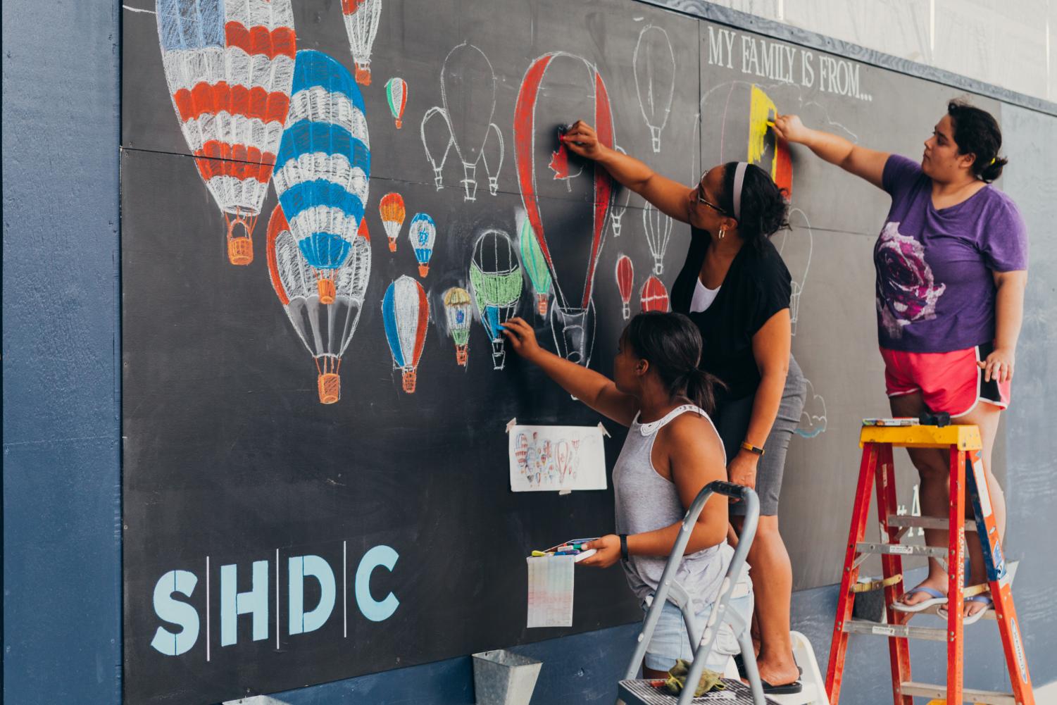 Honors drawing student Alona Miller, assisted by a teacher and student, draws her first place design on the chalk wall in the Van Aken District.
